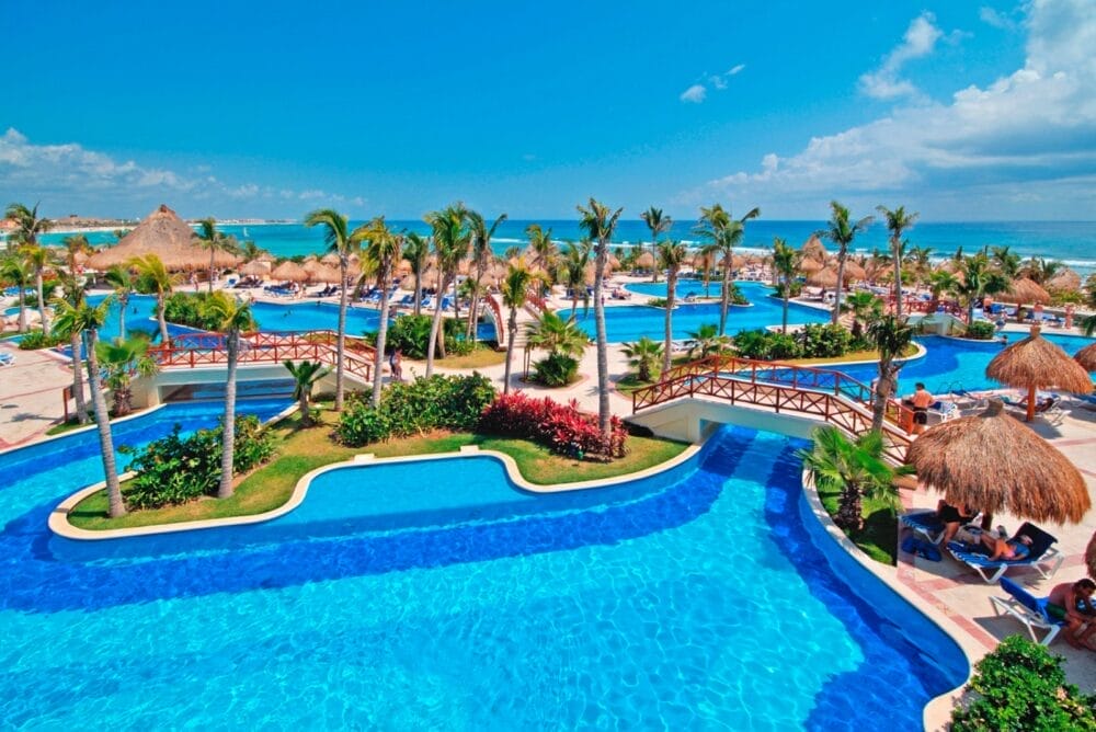 Un resort tropicale ad Akumal con diverse piscine blu, palme, ponti di legno, sedie a sdraio e ombrelloni di paglia, il Bahia Principe Luxury Akumal si affaccia su una spiaggia sabbiosa e sull'oceano sotto un cielo blu brillante.