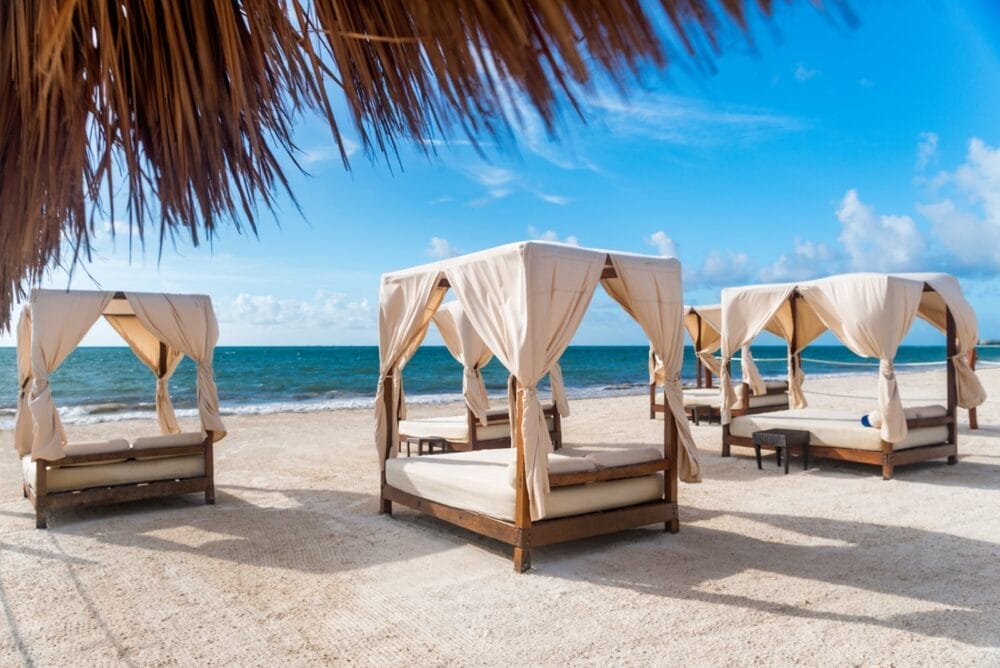 Quattro letti a baldacchino con drappi bianchi attendono sulla spiaggia sabbiosa dell'Ocean Maya Royale Searesort, vicino all'oceano sotto un cielo azzurro e limpido. Un tetto di paglia offre un'ombra parziale in questo sereno paradiso costiero.