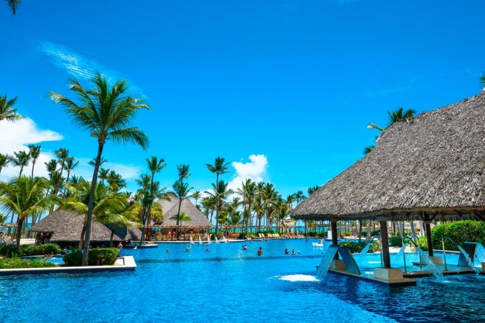 Una piscina tropicale al Barcelo Bavaro Palace con cabine dal tetto di paglia, palme e persone che nuotano sotto un cielo azzurro e limpido: vivete il meglio di Punta Cana in questo resort all-inclusive.