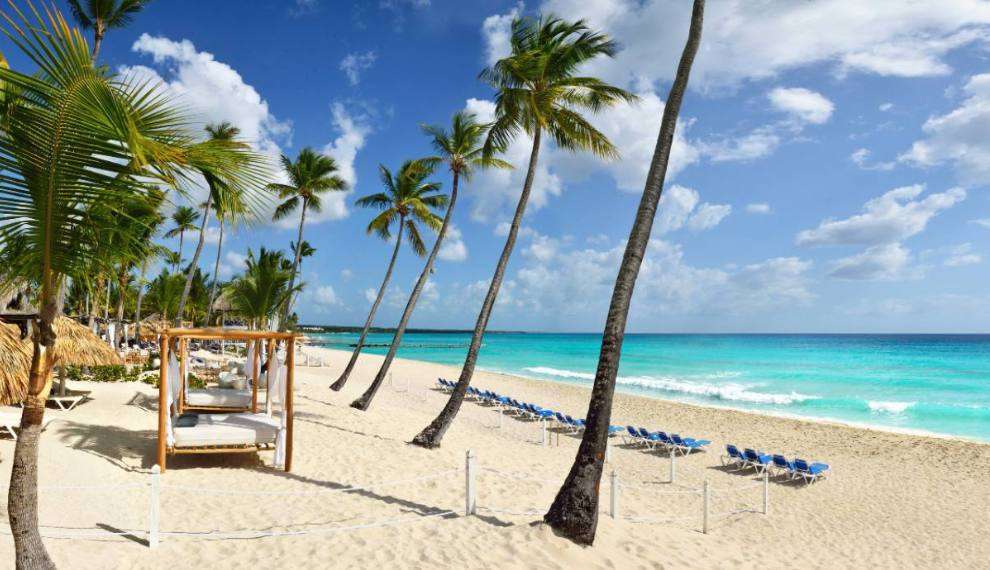 La spiaggia di sabbia bianca del Catalonia Royal La Romana Searesort, con alte palme, cabine ombreggiate e file di sedie a sdraio blu che si affacciano sulle onde turchesi dell'oceano: una fuga per soli adulti sotto un cielo blu brillante con nuvole sparse.