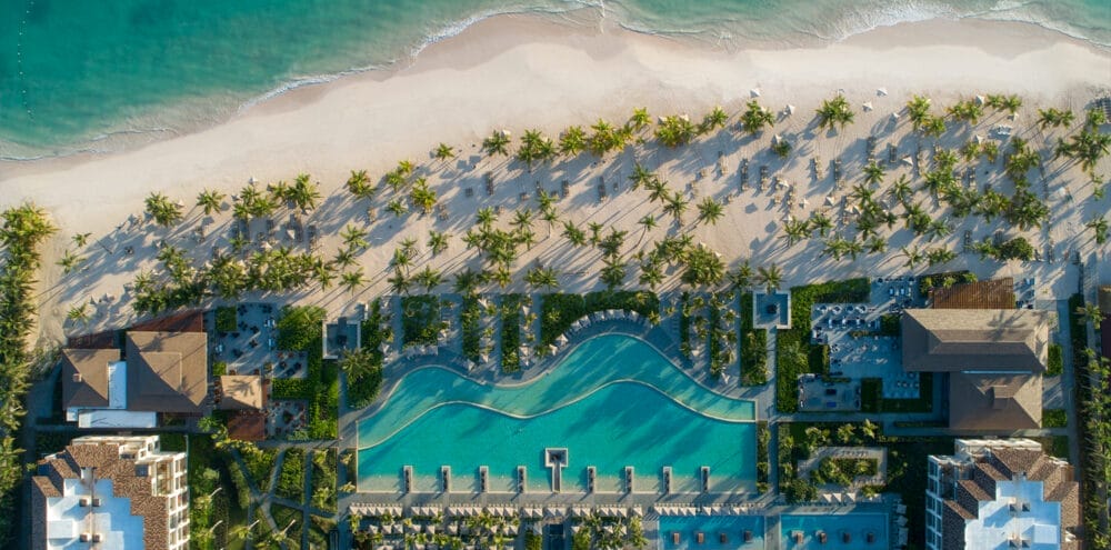 Vista aerea del Lopesan Costa Bavaro, un resort sulla spiaggia con palme, una grande piscina centrale, giardini lussureggianti ed edifici eleganti, tutti affacciati su una spiaggia sabbiosa e sulle calme onde turchesi dell'oceano.