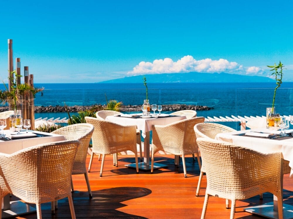 Terrazza ristorante all'aperto con sedie in vimini e tavoli apparecchiati del Seaclub H10 Gran Tinerfe, con vista su un mare azzurro e calmo e sulle montagne lontane sotto un cielo limpido e luminoso.