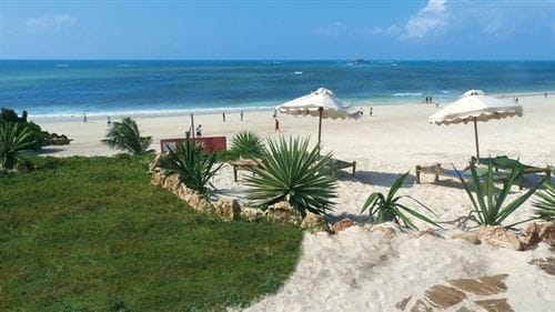 Una spiaggia sabbiosa con ombrelloni bianchi e sedie a sdraio del Bravo Garoda Resort, delimitata da piante ed erba verde, si affaccia sull'oceano blu sotto un cielo luminoso. Sullo sfondo, persone che camminano lungo la riva.