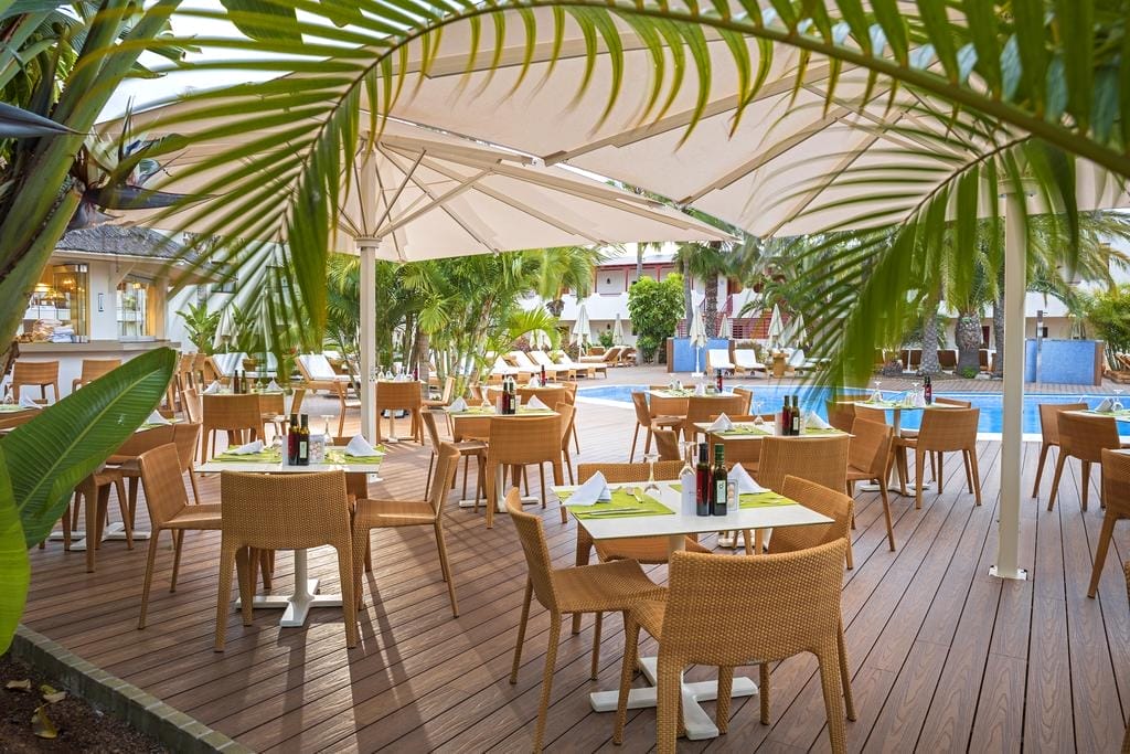 Il ristorante all'aperto dell'Alua Suites Fuerteventura dispone di sedie e tavoli in vimini con tovaglioli e condimenti, sotto grandi ombrelloni, circondati da rigogliose piante tropicali e palme, accanto a una piscina con sedie a sdraio.