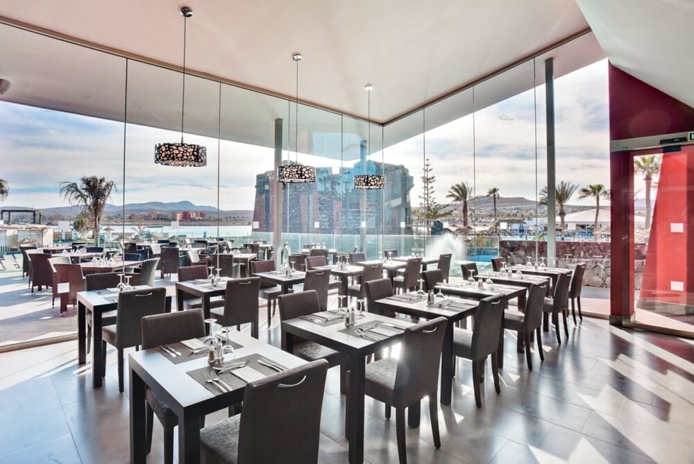 Il moderno ristorante del Seaclub Barcelo Fuerteventura è caratterizzato da ampie vetrate, tavoli e sedie scuri ordinati e una vista panoramica sulle palme, sull'acqua e sugli edifici del Castillo sotto un cielo soleggiato.