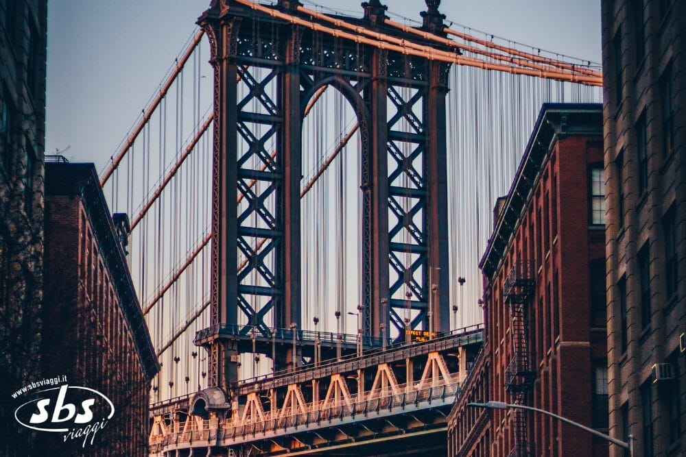 Veduta del Manhattan Bridge incorniciato da edifici in mattoni al tramonto, con luci soffuse che evidenziano la struttura in acciaio e i cavi del ponte. Il logo di SBS Viaggi e la scritta "Bozza automatica" appaiono nell'angolo in basso a sinistra.