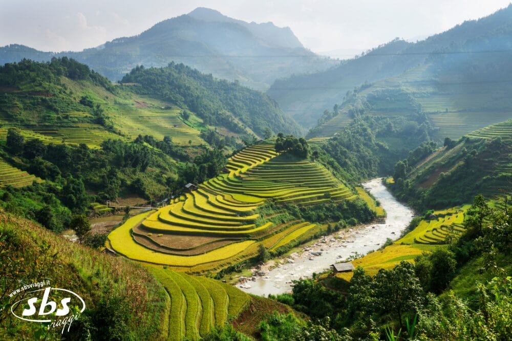 Campi di riso a terrazze si snodano lungo verdi colline accanto a un fiume sinuoso sotto un cielo nebuloso, con le montagne sullo sfondo. La vegetazione rigogliosa e il testo "Bozza automatica" accompagnano il logo "sbs image" nell'angolo in basso a sinistra.