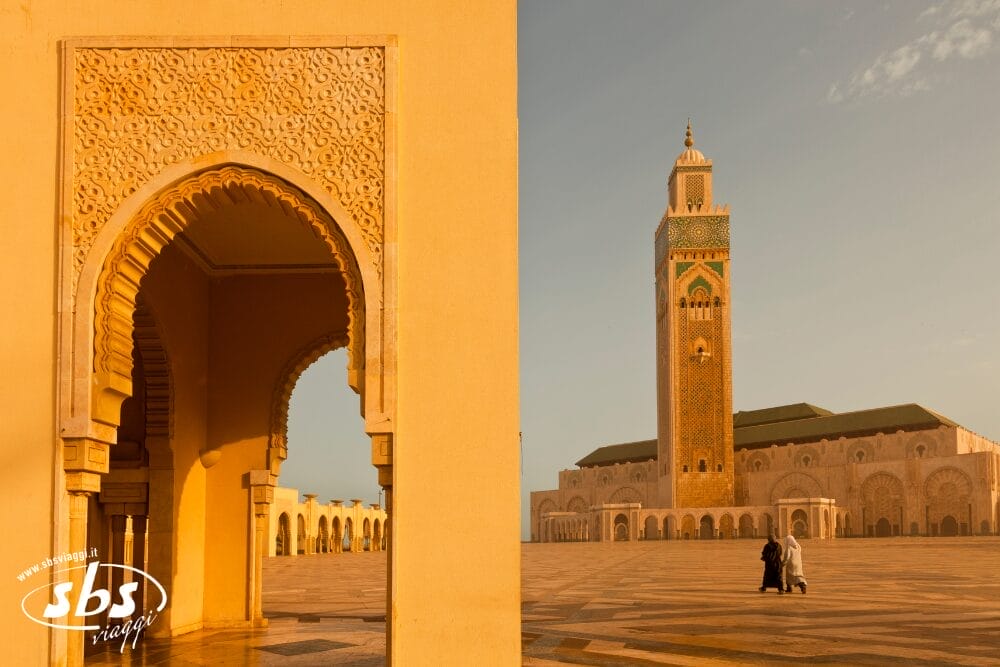 La luce dorata del sole illumina la moschea Hassan II di Casablanca, in Marocco, mettendo in risalto il suo minareto ornato. Due persone attraversano il vasto cortile vuoto incorniciato da un arco decorativo, una scena di tranquillità quasi automatica.