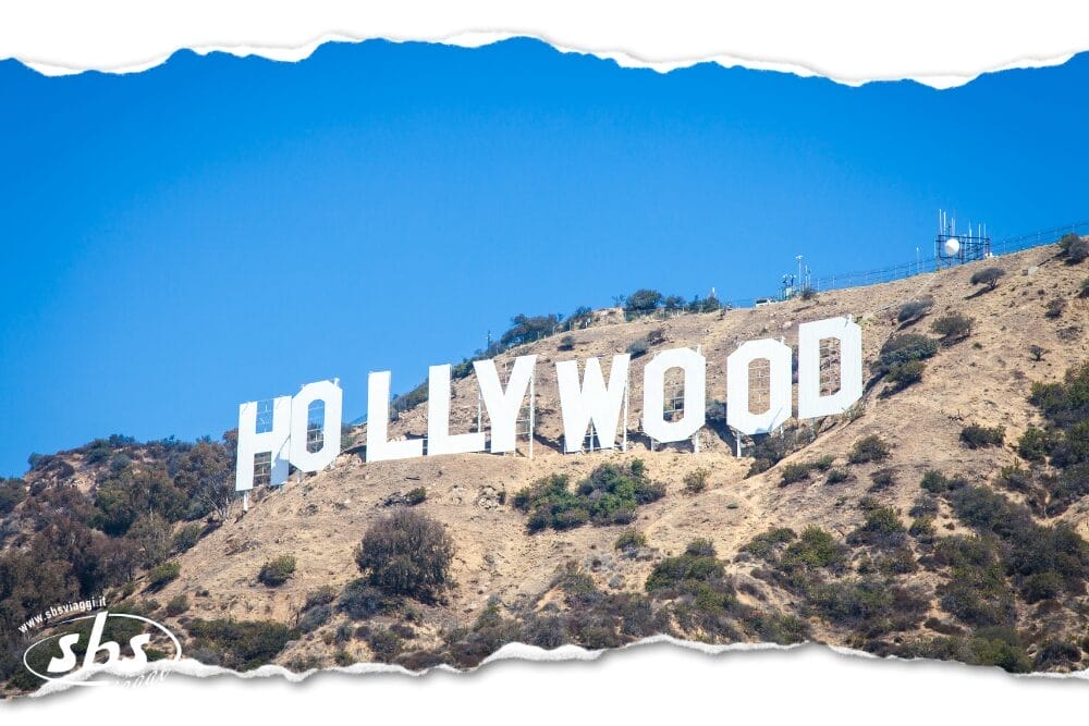 La famosa insegna bianca di Hollywood si erge su una collina secca e cespugliosa sotto un cielo azzurro e limpido, con un effetto di carta strappata e la scritta "Bozza automatica" accanto al piccolo logo "sbs" nell'angolo in basso a sinistra.
