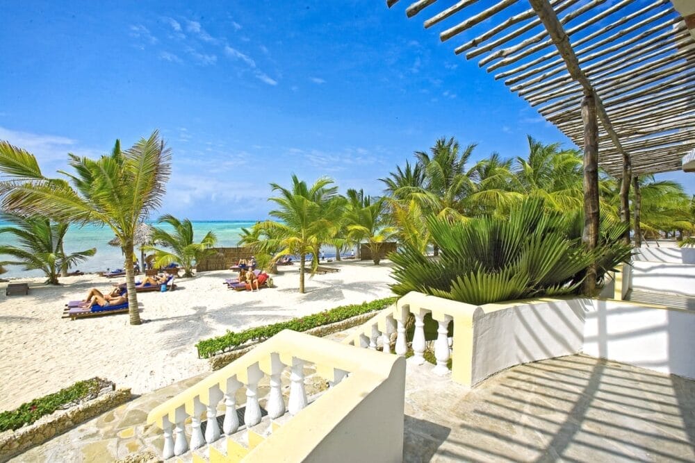 Una scena di sole sulla spiaggia di Bravo Watamu con palme, persone sdraiate sulle sedie, sabbia bianca, acqua turchese e una terrazza ombreggiata con ringhiere bianche in primo piano.