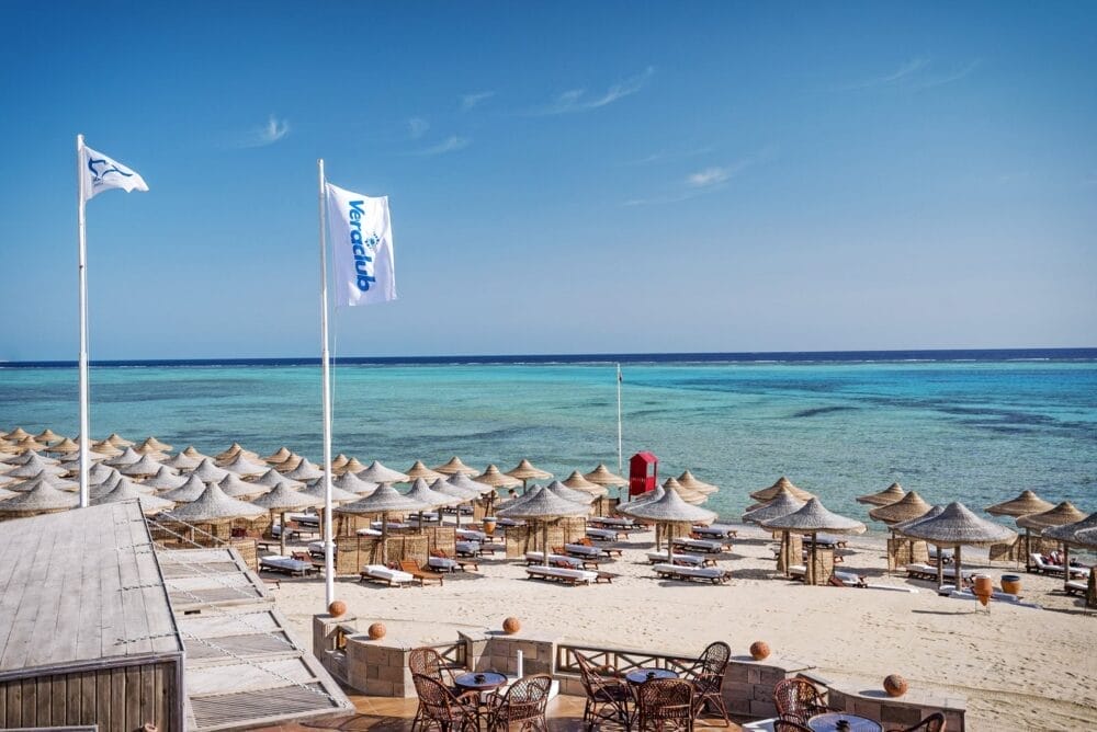 Uno stabilimento balneare con file di lettini e ombrelloni di paglia sulla sabbia bianca, affacciati su un mare azzurro e limpido. Al Veraclub Emerald Lagoon, le bandiere bianche sventolano nella brezza e una stazione di salvataggio rossa si trova vicino alla riva, sotto un cielo limpido e soleggiato.
