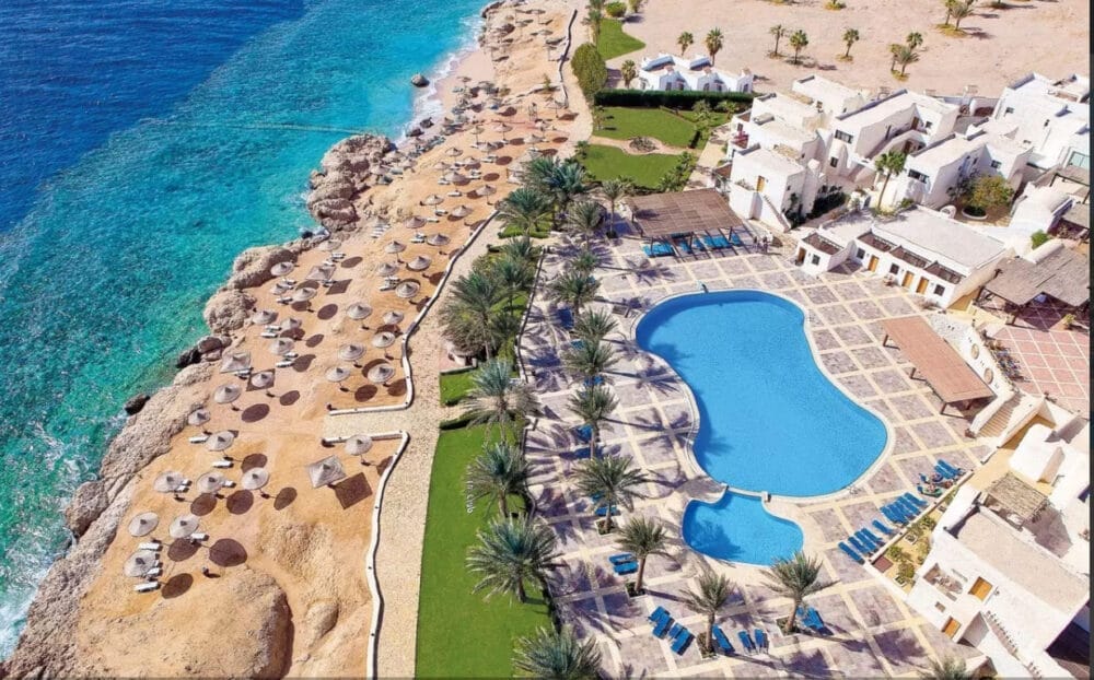 Vista aerea del resort Veraclub Sharm a Sharm El Sheikh, caratterizzato da una grande piscina sinuosa, palme, sedie a sdraio ed edifici bianchi accanto a una costa rocciosa e a un mare turchese con ombrelloni e sedie a sdraio lungo la riva.