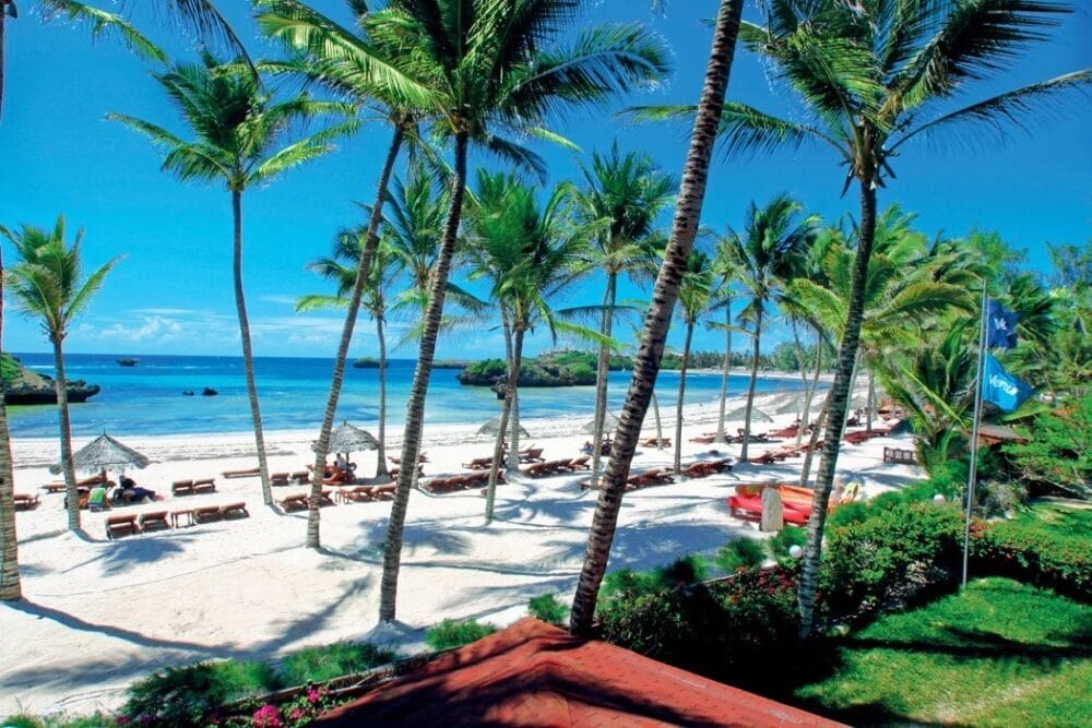 Le palme costeggiano la spiaggia di sabbia bianca del Veraclub Crystal Bay, dove sedie a sdraio e ombrelloni invitano al relax. Le persone si rilassano in riva all'oceano turchese sotto un cielo limpido, mentre un fogliame lussureggiante delimita questo paradiso tropicale.