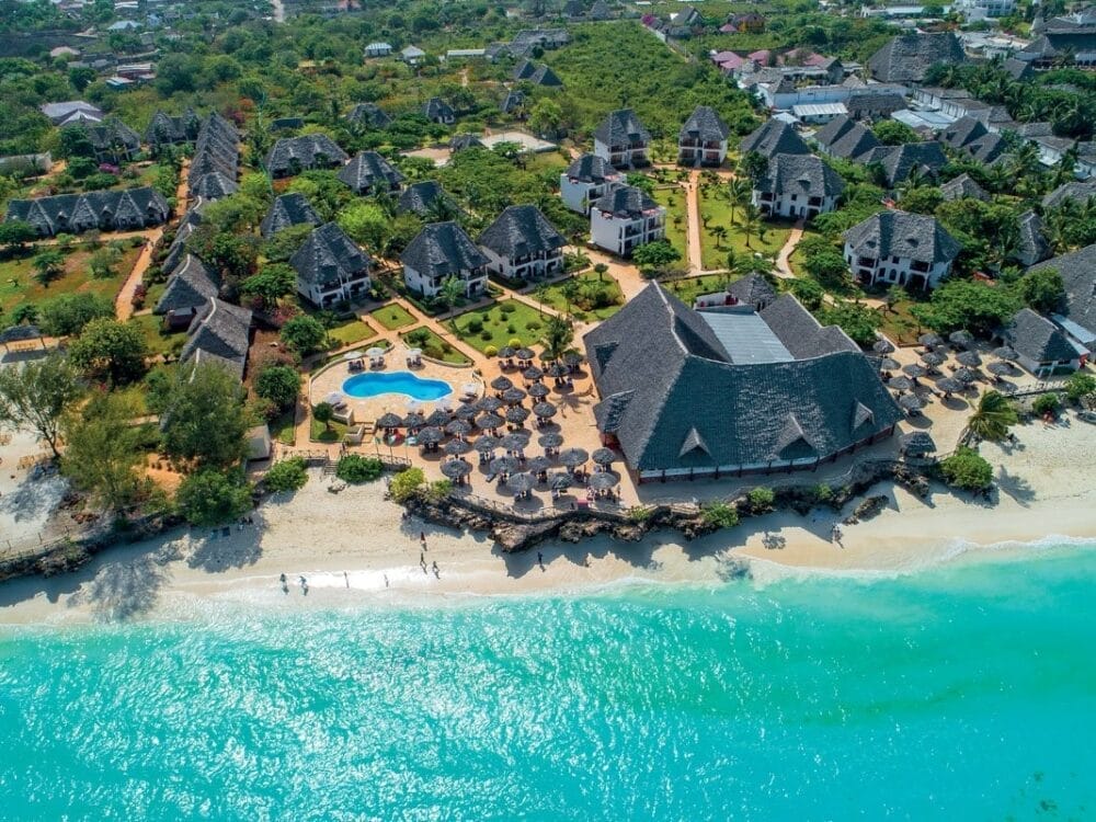Vista aerea del resort Veraclub Sunset Beach con edifici dai tetti di paglia, una piscina, lettini con ombrelloni e persone che passeggiano su una spiaggia di sabbia bianca in riva al mare turchese. Il verde lussureggiante circonda l'area.