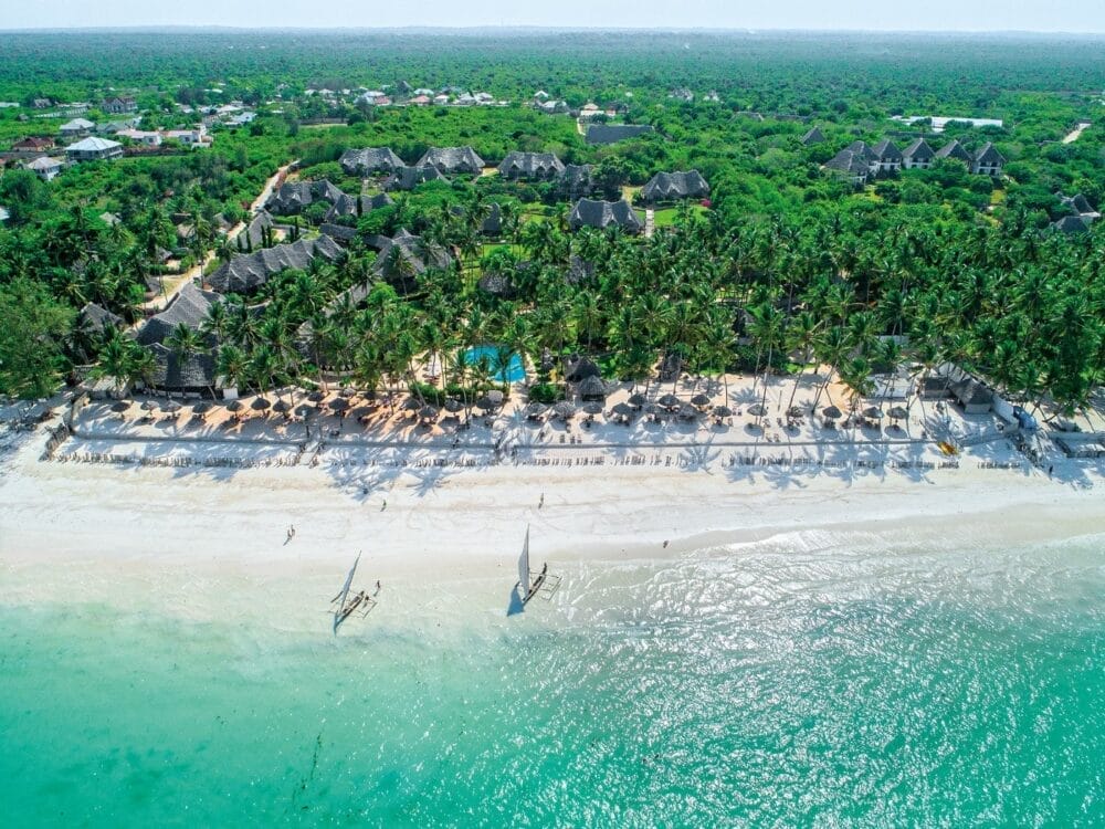 Veduta aerea di Veraclub Zanzibar, un resort tropicale sulla spiaggia con edifici dai tetti di paglia, palme, spiaggia di sabbia bianca, acqua turchese e due barche a vela vicino alla riva. Una fitta vegetazione circonda il tranquillo villaggio di Zanzibar.