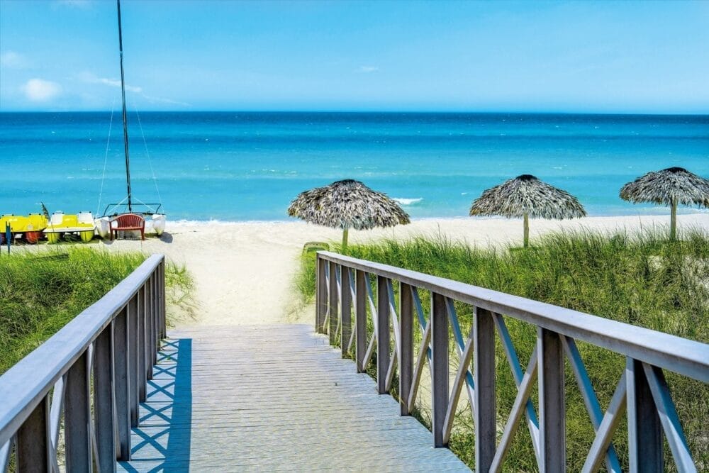 Una passerella di legno conduce alla spiaggia sabbiosa del Veraclub Starfish Varadero, dove ombrelloni di paglia, erba verde e calme onde oceaniche blu attendono sotto un cielo limpido. Piccole barche colorate vanno alla deriva sulla sinistra di questo invitante hotel di Cuba.