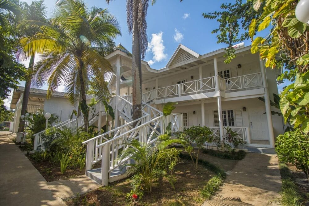 L'edificio tropicale a due piani del Veraclub Negril è luminoso e bianco, con balconi e ringhiere, circondato da palme verdi e lussureggianti sotto un cielo blu con nuvole sparse: una fuga perfetta in questo resort di Negril in Giamaica. Le scale conducono agli ingressi principali.