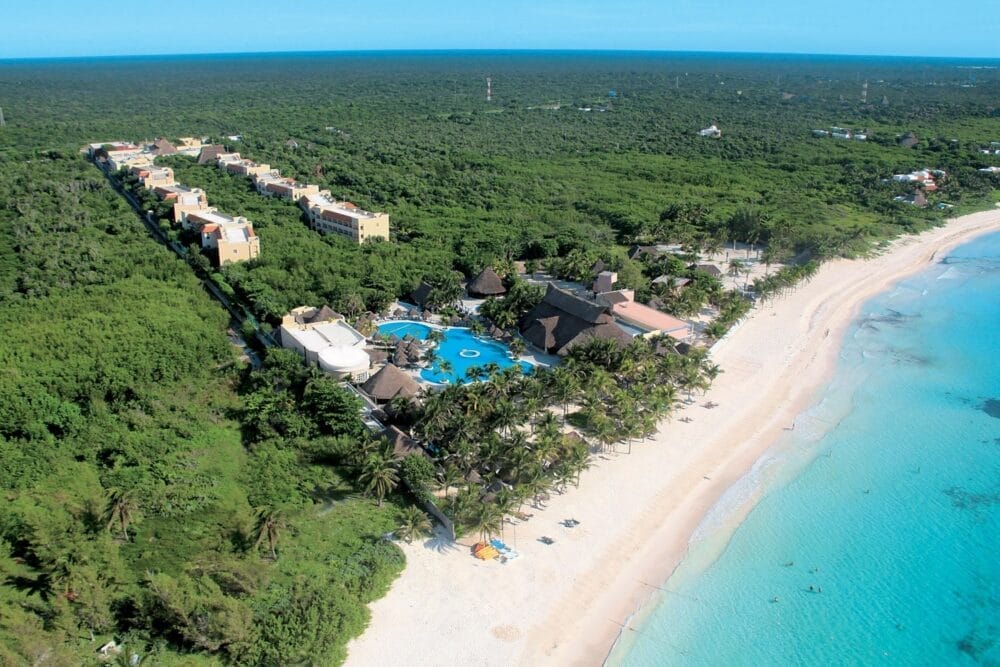 Vista aerea del Veraclub Royal Tulum, un resort all-inclusive in Messico immerso in una fitta foresta verde, con piscine, palme e una spiaggia di sabbia bianca accanto a un'acqua turchese e trasparente. File di edifici si estendono nell'entroterra tra gli alberi.