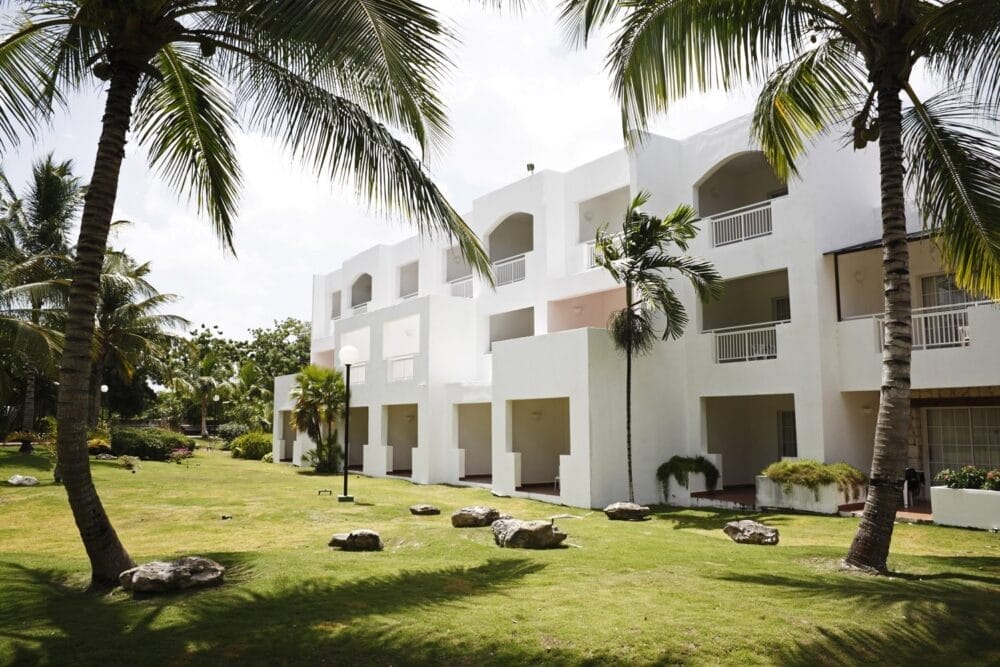 Un edificio bianco a tre piani con balconi è circondato da erba verde, palme e rocce al Sunscape Dominicus, sotto un cielo parzialmente nuvoloso in un lussureggiante ambiente tropicale.