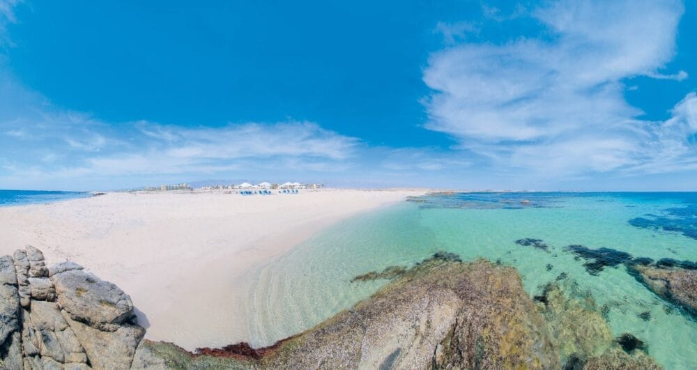 Una vista panoramica di una spiaggia sabbiosa con acqua limpida e turchese, rocce in primo piano e un cielo blu con nuvole sparse. In lontananza sono visibili gli ombrelloni bianchi e le sedie a sdraio del resort Veraclub Salalah.