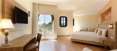 Spaziosa e moderna camera d'albergo dell'Iberostar Palmeraie Marrakech con ampio letto, scrivania, TV a parete e porta scorrevole in vetro su un balcone con posti a sedere all'aperto. La luce naturale riempie la stanza, perfetta per un soggiorno rilassante al Kappa Club.