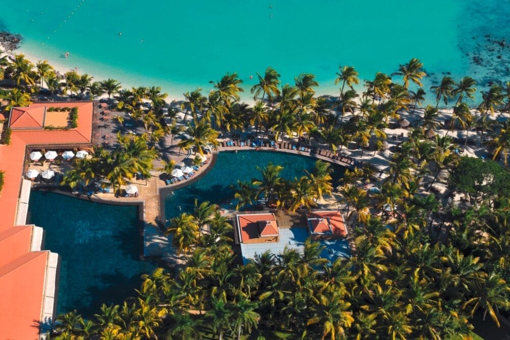 Vista aerea del resort Mauricia Beachcomber con mare turchese, spiaggia sabbiosa, palme, piscine ed edifici dai tetti rossi circondati da una vegetazione lussureggiante.