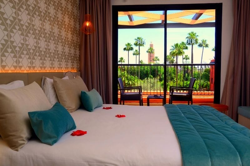 Un'accogliente camera dell'hotel Kappa City con un letto ben fatto, cuscini beige e verde acqua e petali di rosa. Le porte scorrevoli in vetro si aprono su un balcone con due sedie, con vista sulle palme e sui soleggiati Giardini della Medina.