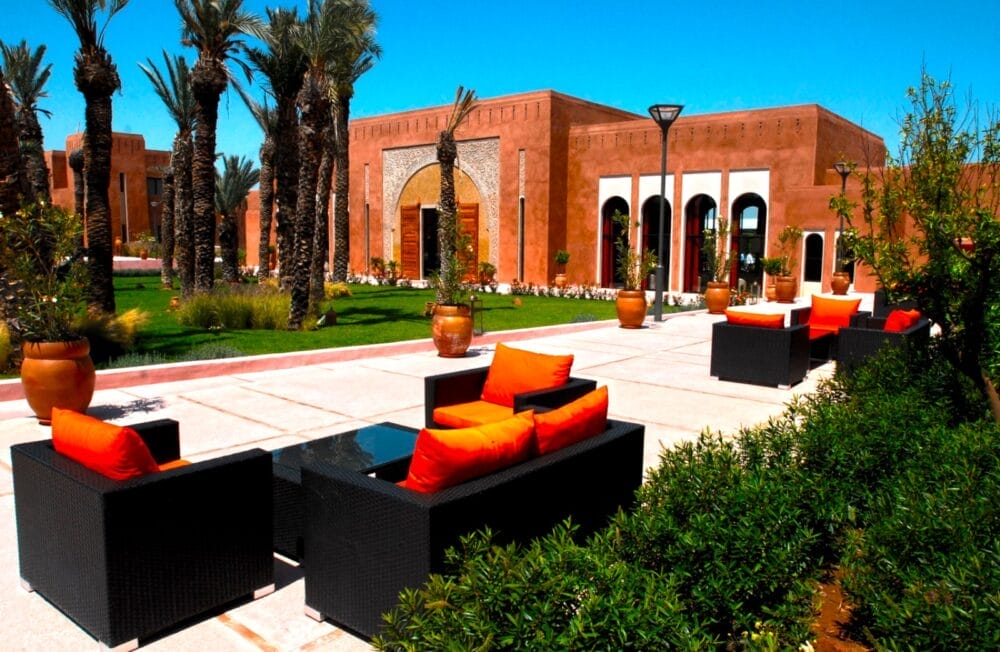 Area salotto all'aperto con divani in vimini neri e cuscini arancioni brillanti, di fronte all'elegante edificio Kenzi con archi. Palme e verde circondano il patio sotto un cielo azzurro, evocando il fascino di Agdal e Medina.