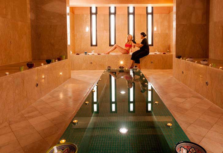 Una serena sala termale dell'Aqua Mirage Marrakech presenta pareti in marmo marrone, una piscina centrale rettangolare e candele. Una donna in accappatoio siede a bordo piscina mentre un membro del personale si prende cura di lei, con luci soffuse che creano un'atmosfera rilassante.