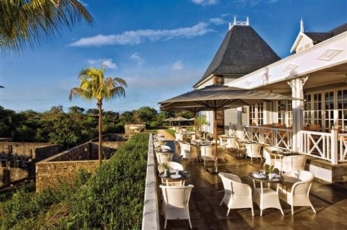 Terrazza all'aperto di un ristorante del Maritim Resort, con sedie e tavoli in vimini bianchi all'ombra di grandi ombrelloni, con vista sul verde, sulle palme e sui muri storici in pietra sotto il cielo azzurro di Mauritius.