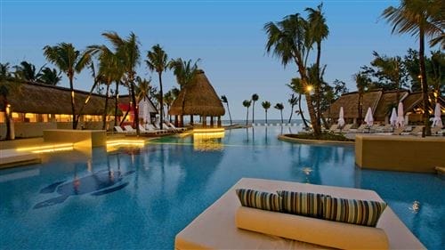 Una lussuosa piscina tropicale al tramonto dell'Ambre Sun Resort, a Mauritius, circondata da palme, edifici con tetti di paglia, sedie a sdraio e ombrelloni, con un'atmosfera tranquilla e l'oceano visibile sullo sfondo.