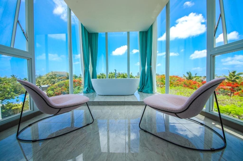 La moderna camera dell'Ocean V Hotel è caratterizzata da due sedie rosa che si affacciano su una vasca da bagno freestanding bianca e da finestre a tutta altezza che offrono una vista panoramica sulla vegetazione lussureggiante e su un cielo blu brillante con nuvole.