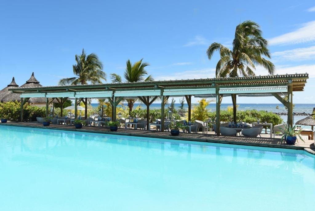 Il Veranda Pointe Aux Biches di Mauritius è un resort sulla spiaggia tropicale con una piscina azzurra, posti a sedere all'ombra sotto un pergolato di legno, palme e l'oceano che scintilla sullo sfondo sotto un cielo soleggiato.