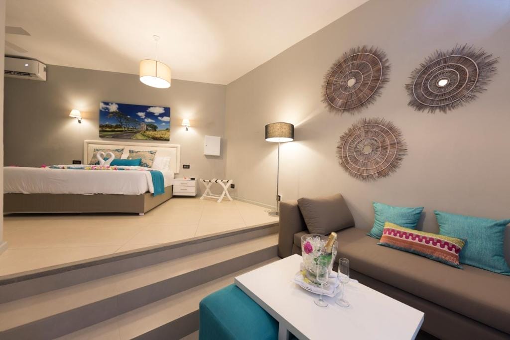 Una moderna camera dell'hotel Azur Paradise con un letto e cuscini colorati, un divano con cuscini blu e rosa, un tavolino bianco con fiori e bevande e opere d'arte decorative su pareti grigio chiaro. Lo spazio è ben illuminato e invitante.