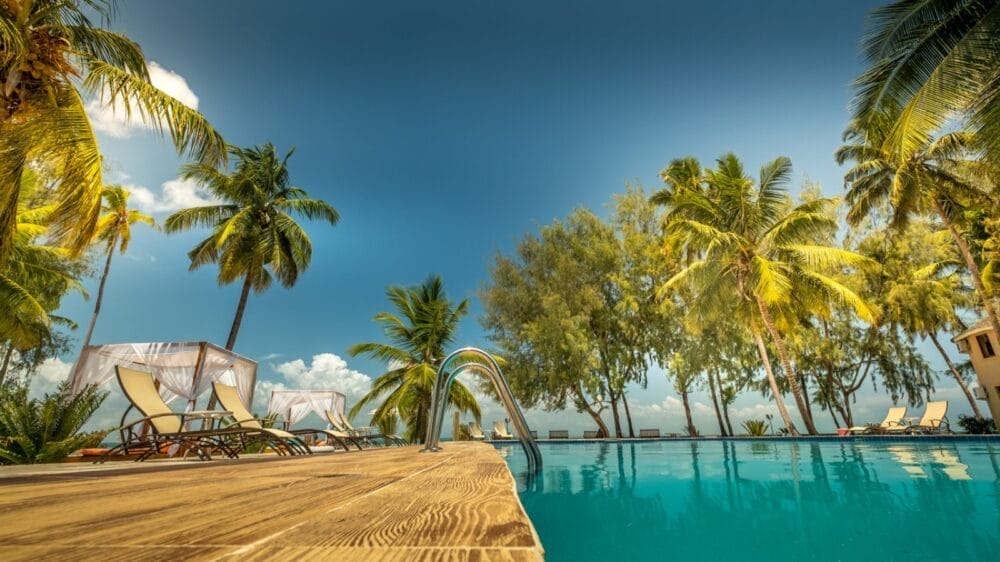 Una lussuosa scena a bordo piscina al Kae Beach Zanzibar con sedie a sdraio, lettini sotto baldacchini bianchi, palme e un cielo limpido e azzurro che si riflette nella piscina, evocando una rilassante atmosfera da resort tropicale.