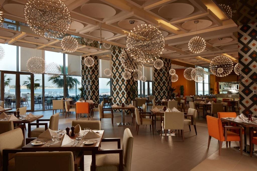 I moderni interni del ristorante del Royal M Hotel Abu Dhabi sono caratterizzati da pareti a motivi geometrici, lampadari rotondi, tavoli ordinati e grandi finestre con vista sulle palme e sull'oceano. Alcune sedie sono arancioni, altre beige.
