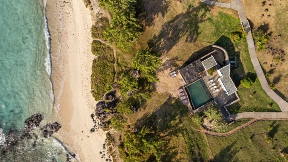 Vista aerea di una casa moderna con piscina vicino a una spiaggia sabbiosa a Constance Tekoma Rodrigues. La casa è circondata da verde e sentieri, con l'acqua turchese dell'oceano e alcune rocce visibili sulla riva.
