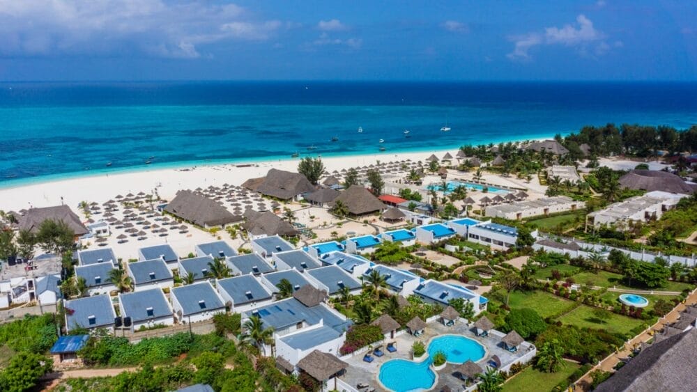 Vista aerea del Bravo Kendwa Beach Resort, caratterizzato da una spiaggia di sabbia bianca, un oceano turchese, diverse piscine, palme, edifici con tetti di paglia e sedie a sdraio disposte lungo la riva sotto un cielo azzurro e soleggiato.