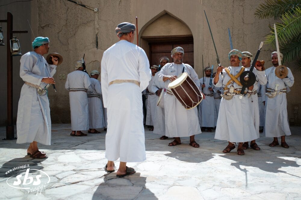 Un gruppo di uomini in abiti tradizionali omaniti esegue una danza culturale con spade e tamburi in un cortile all'aperto, mentre un uomo guida l'esibizione ispirata all'automatica Bozza.