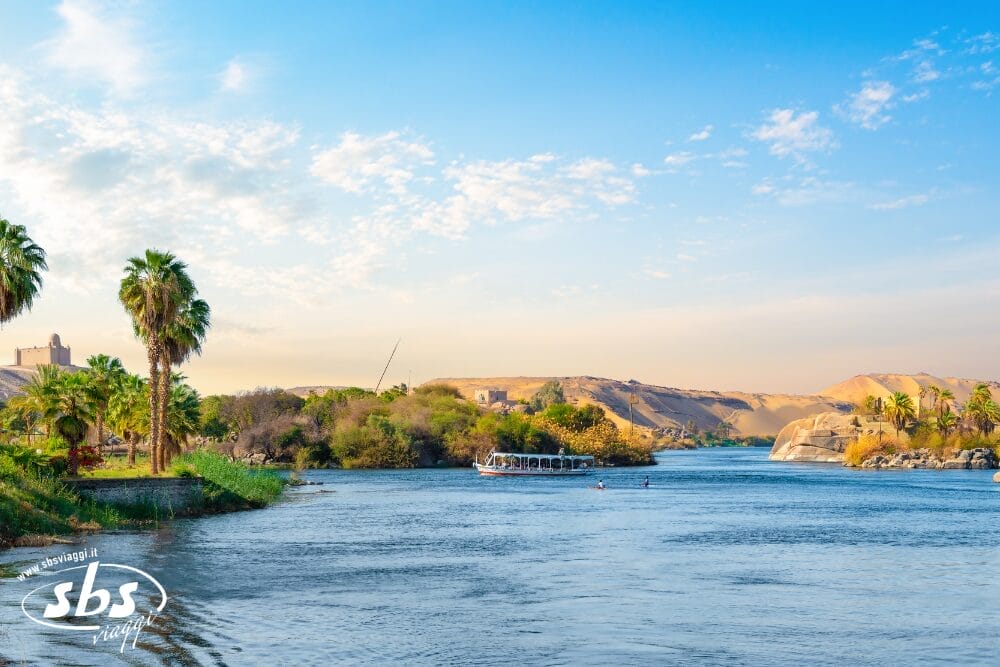 Un fiume panoramico scorre tra palme lussureggianti e colline sabbiose sotto un cielo blu brillante. Una barca bianca con passeggeri si muove sull'acqua, mentre il verde e le rocce costeggiano le sponde del fiume in questo paesaggio automatico di Bozza.