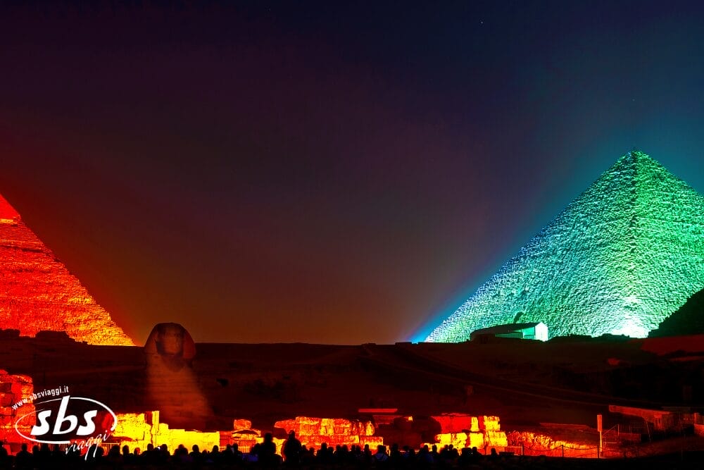 Le Piramidi di Giza e la Sfinge illuminate di notte con luci colorate - rosse, verdi e blu - contro un cielo scuro, con una folla di persone in primo piano che osserva lo spettacolo automatico di Bozza.