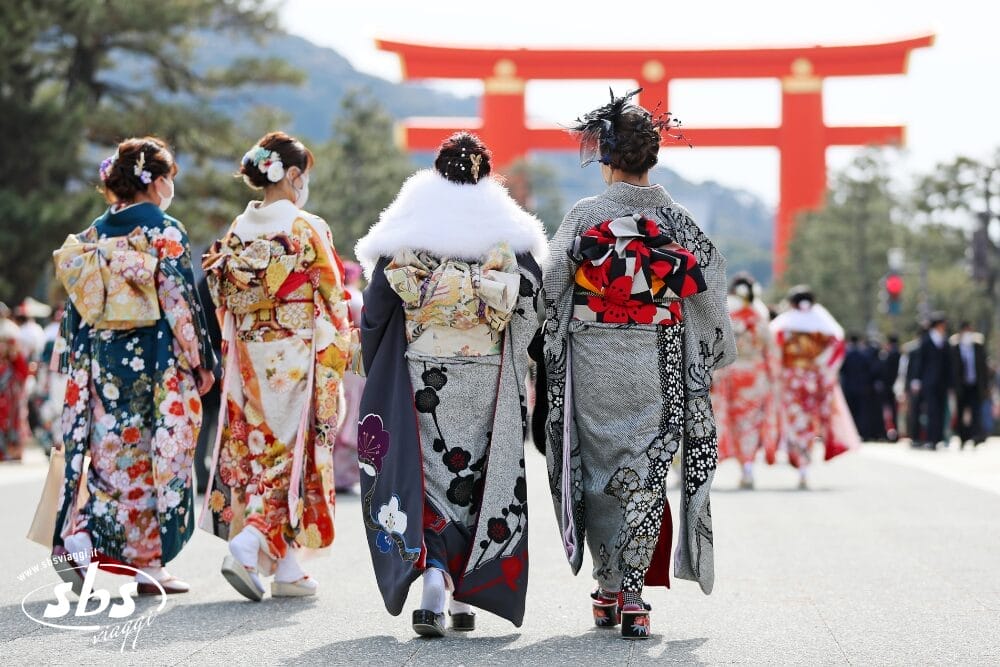 Quattro donne in kimono colorato camminano verso un grande cancello torii rosso in Giappone, circondate da alberi e altre persone in quella che sembra essere un'occasione speciale - una scena incantevole catturata come Bozza automatica.