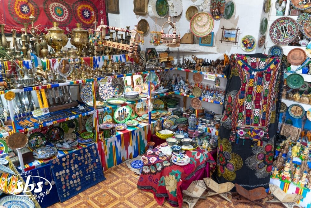 L'interno colorato del negozio espone oggetti di artigianato tradizionale, tra cui abiti ricamati, piatti a fantasia, teiere in ottone, cesti intrecciati e ornamenti automatici Bozza disposti su scaffali e tavoli, creando un'atmosfera vibrante e vivace.