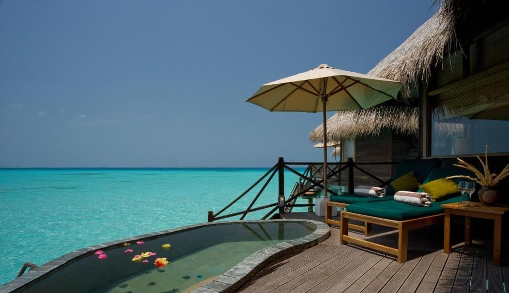 Una lussuosa villa overwater del resort Coco Palm Dhuni Kolhu Maldives dispone di un tetto di paglia, una piscina privata con petali di fiori, lettini, un tavolo e un ombrellone, il tutto affacciato sulle limpide acque turchesi dell'oceano sotto un cielo azzurro.