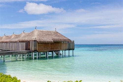 Bungalow overwater con tetto in paglia su palafitte sopra il limpido oceano turchese del Constance Moofushi, un resort di lusso alle Maldive, con la costa sabbiosa e la vegetazione lussureggiante in primo piano sotto un cielo azzurro parzialmente nuvoloso.