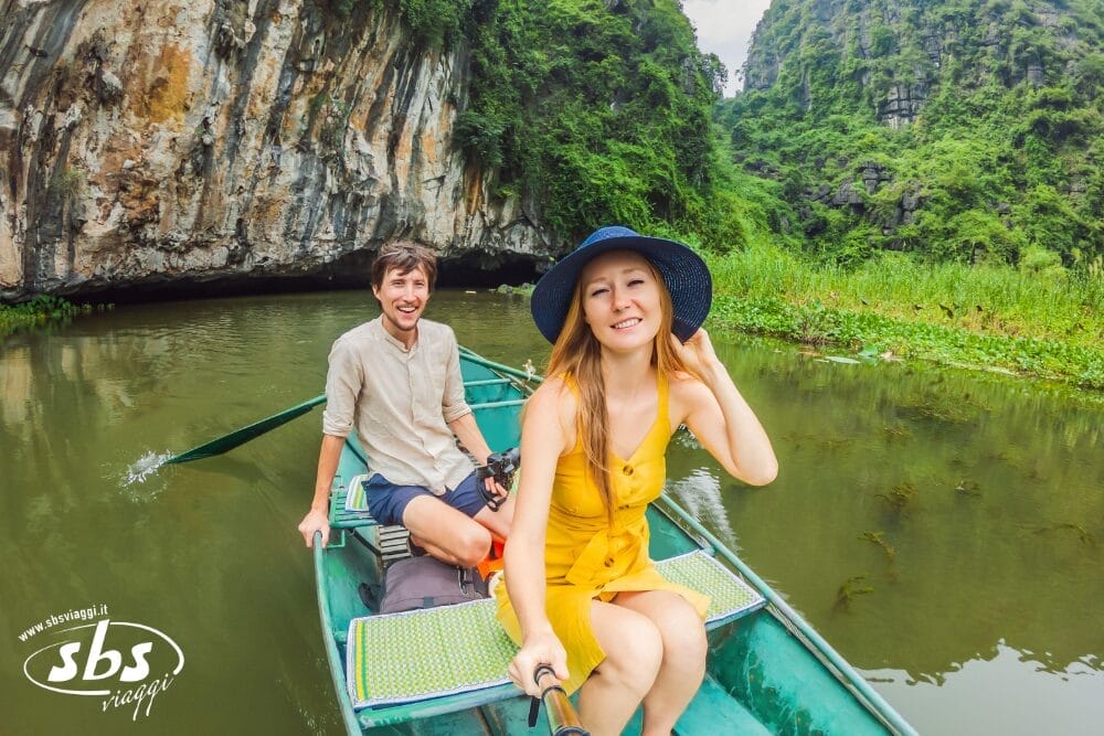 Una coppia sorridente si scatta un selfie mentre viaggia su una piccola barca verde attraverso un canyon fluviale lussureggiante e roccioso. La donna in abito giallo e cappello blu cattura il momento, creando una perfetta Bozza automatica mentre il verde e le scogliere li circondano.