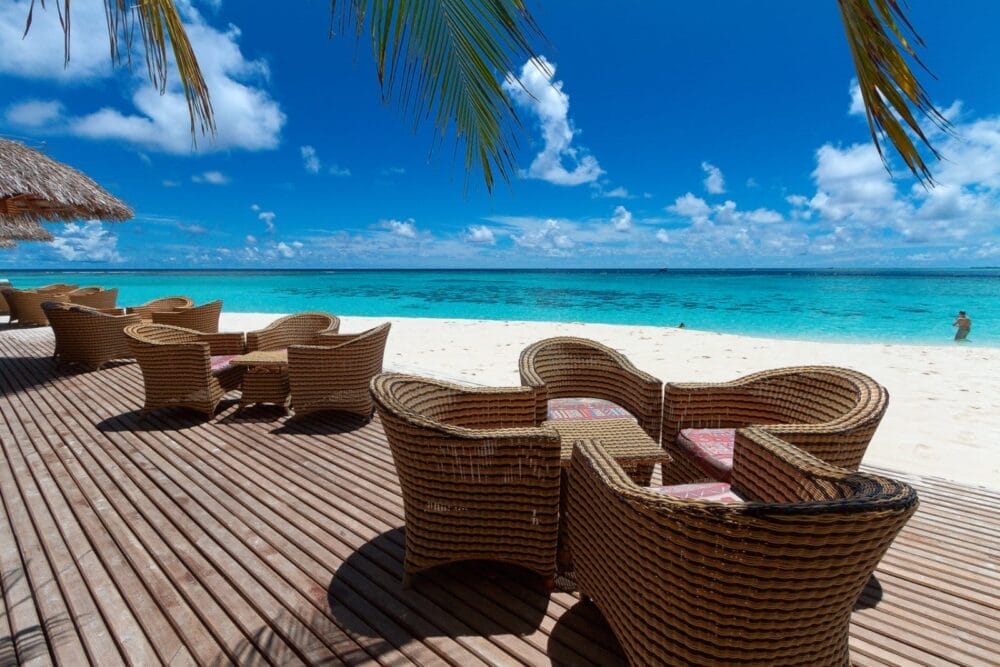 Sedie e tavoli di vimini sono disposti su una terrazza di legno sotto le foglie di palma del Bravo Alimatha', con vista su una spiaggia di sabbia bianca e sull'oceano turchese sotto un cielo blu brillante punteggiato di nuvole.