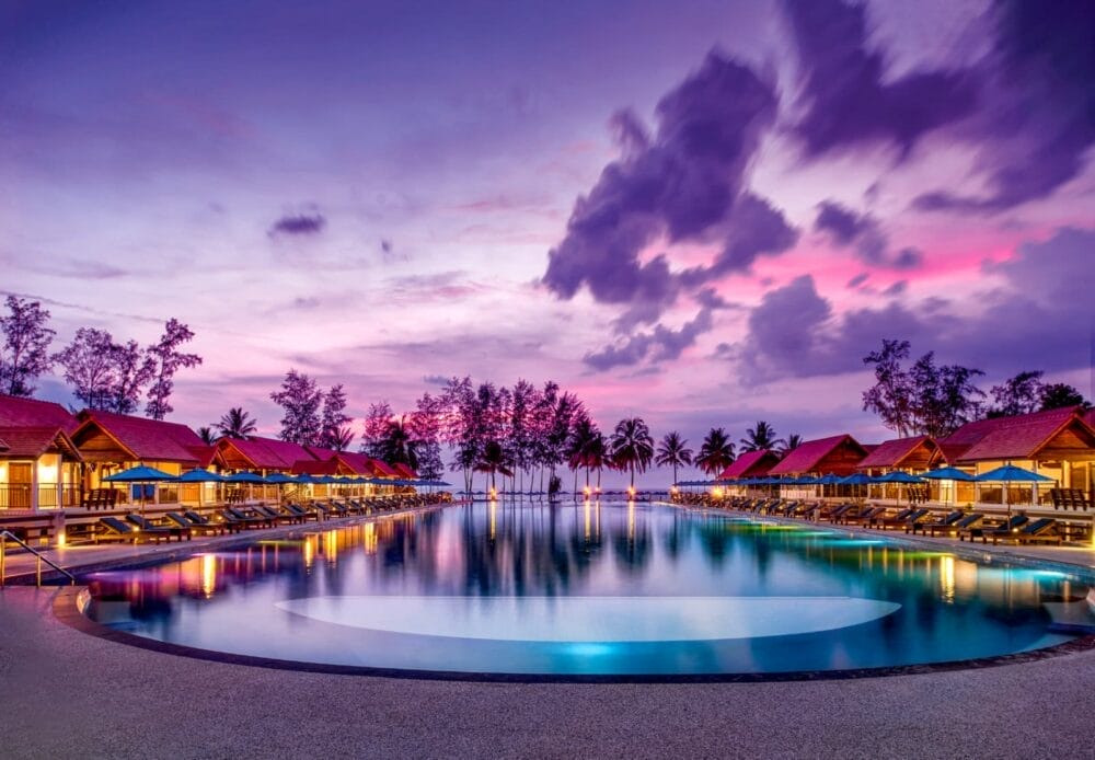La serena piscina a sfioro del Bravo Premium Khao Lak è circondata da sedie a sdraio e bungalow in legno sotto un vibrante cielo viola e rosa al tramonto, con le palme che si stagliano sullo sfondo vicino alla riva.