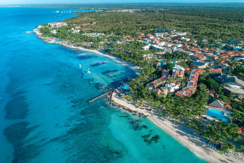 Veduta aerea di una città costiera con acque turchesi, spiagge sabbiose e palme nei pressi della spiaggia Bravo Viva Dominicus, dove edifici dai tetti rossi costeggiano il litorale delimitato da una rigogliosa vegetazione e dalla foresta.