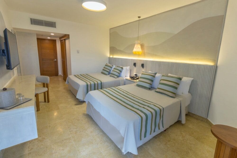 Moderna camera d'albergo Bravo Premium con due letti matrimoniali, ciascuno con biancheria da letto a righe verdi. Lo spazio è caratterizzato da pareti e pavimenti chiari, TV a parete, zona scrivania, luce a sospensione e porte in legno sullo sfondo.